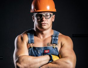 Portrait of a muscular construction worker with arms crossed, wearing goggles and a hard hat.